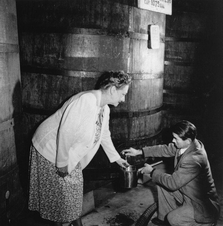 A older woman leaning forward and holding a small metal container in front of a massive wine barrel and a man crouched, pouring wine from the barrel into the container..