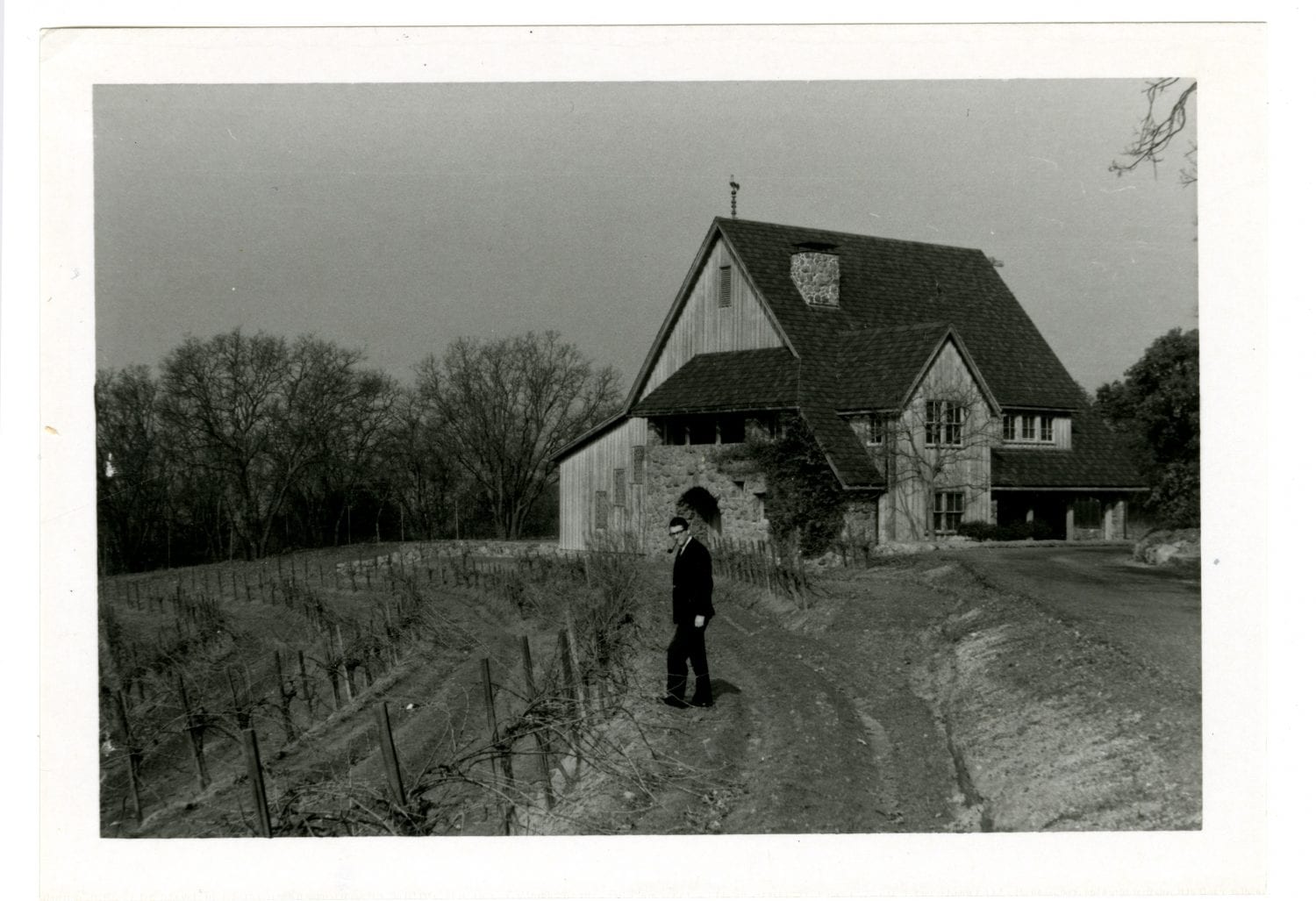 Black and white image of a man in a dark suit, standing next to a vineyard with a winery and trees behind him.