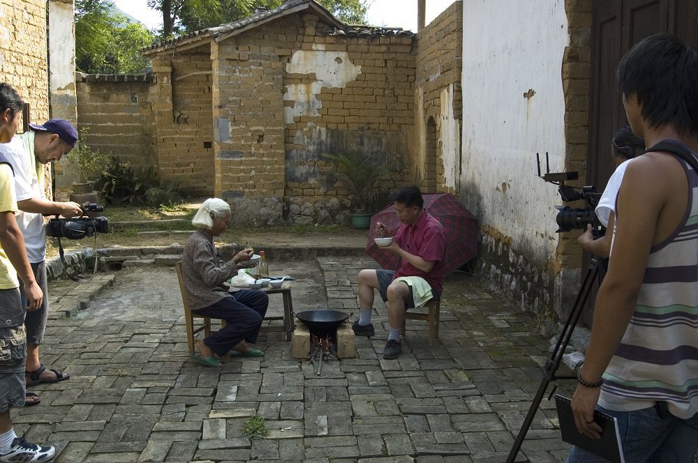Martin Yan with a local woman, a traditional wok between them. Cameras in the foreground film the scene.