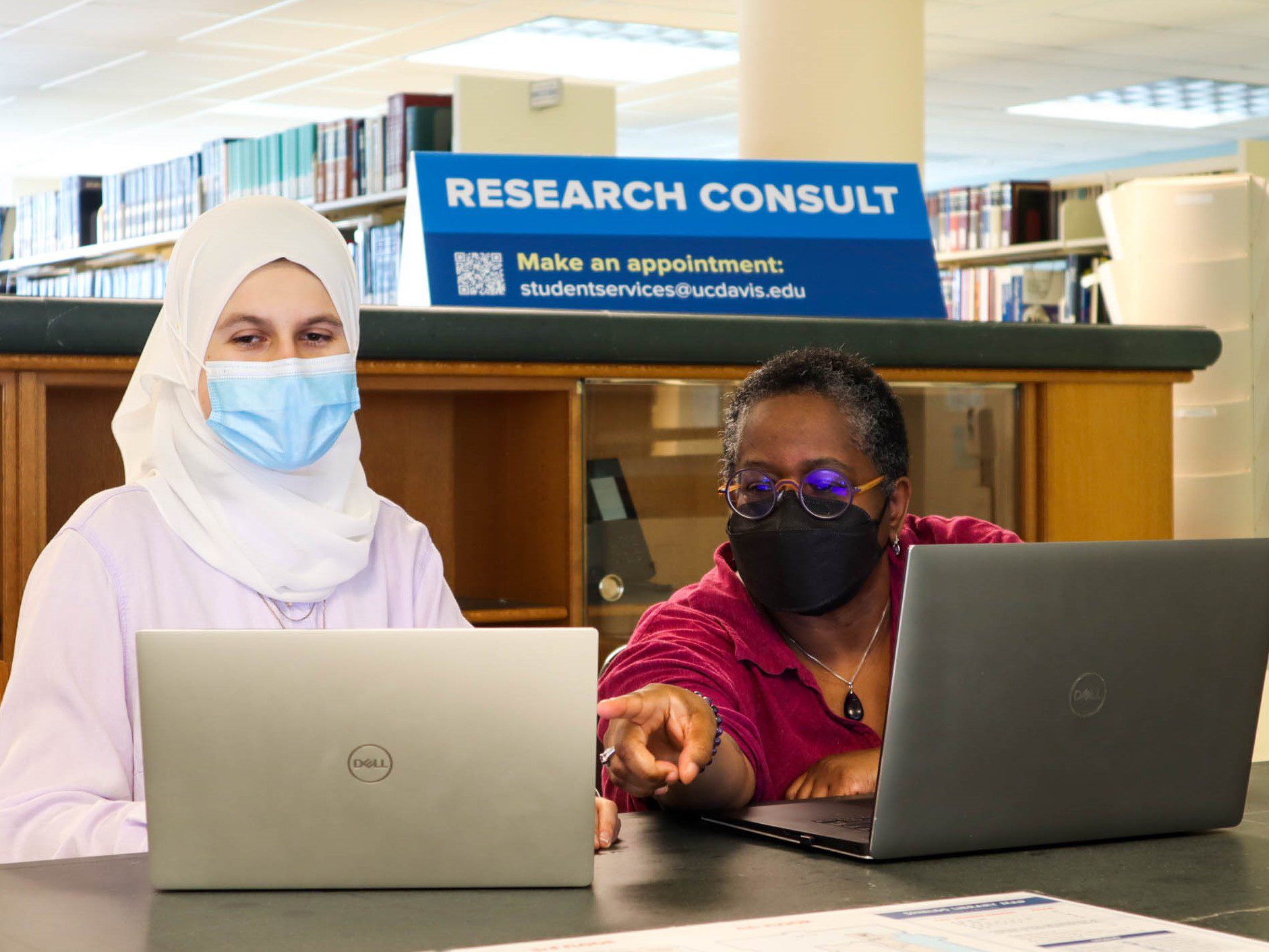 Student looks on as librarian Nancy Wallace points out a library resource on her laptop computer. Sign behind them says Research Consult.