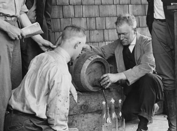 1918 black and white image of Viticulture and Enology Professor William Cruess crouching next to an oak barrel, pouring wine, while students look on.