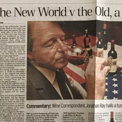 Newspaper clipping of Steven Spurrier sniffing a glass of red wine with a table of wine bottles beside him.