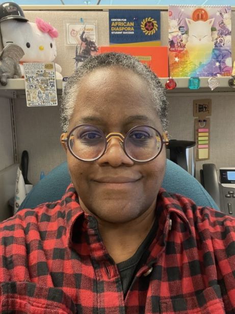 Nancy Wallace wearing a red-and-black shirt and round glasses, sitting at their desk.