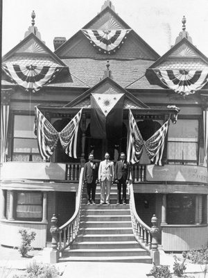 Archival image of Victorian home with three men standing on staircase leading up to front door and US and Phillipines flags.