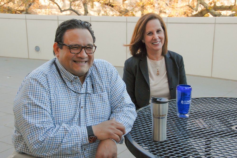 Librarians Roberto Delgadillo and Belen Fernandez sit at a picnic table with cups of coffee