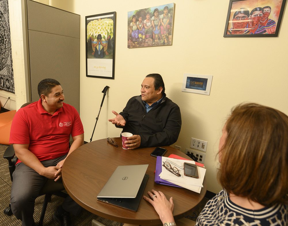 Two men sit at a table smiling and talking, with Chicanx art on the walls behind them