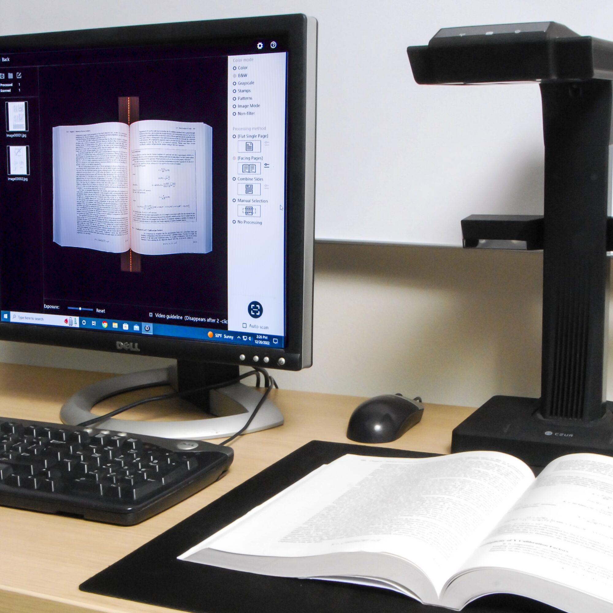 A book lays open on a desk with a black scanner positioned above it. To the left is a computer mouse, keyboard and a monitor displaying an image of the book being scanned for digitization.