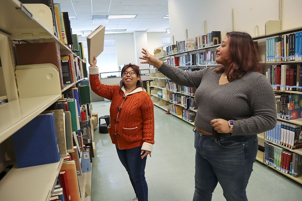 Karis Chun selecting a book from the top shelf of a book stack, while Emily Gavidia reaches out to assist her.