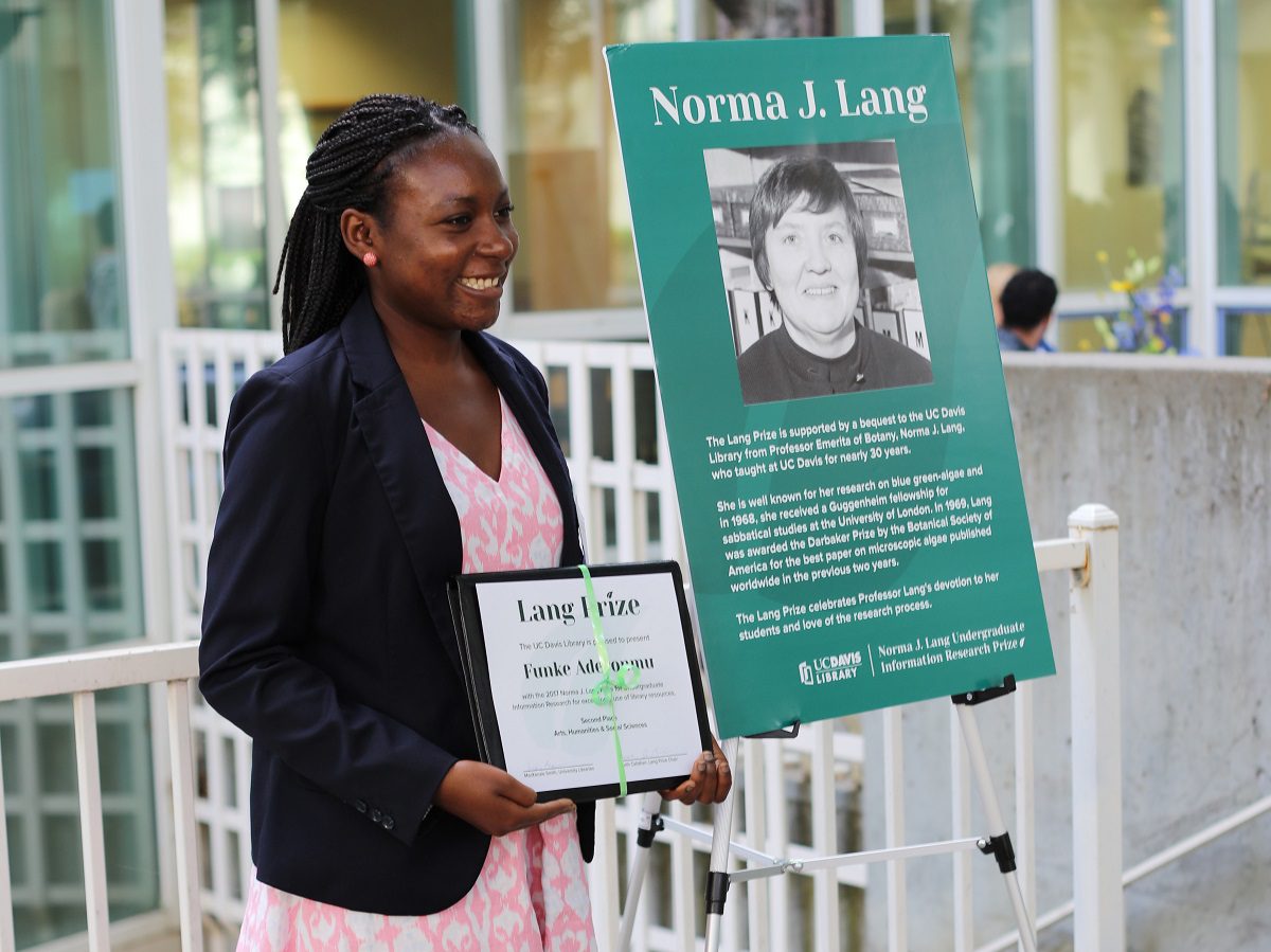 Lang Prize winner in pink dress and black jacket holding a certificate stands next to a poster of Norma J. Lang.