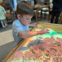 A child shapes sand in the Augmented Reality Sandbox, simulating rain fall and lava flows, as red, green, yellow and blue lights flood his palms and the sand.