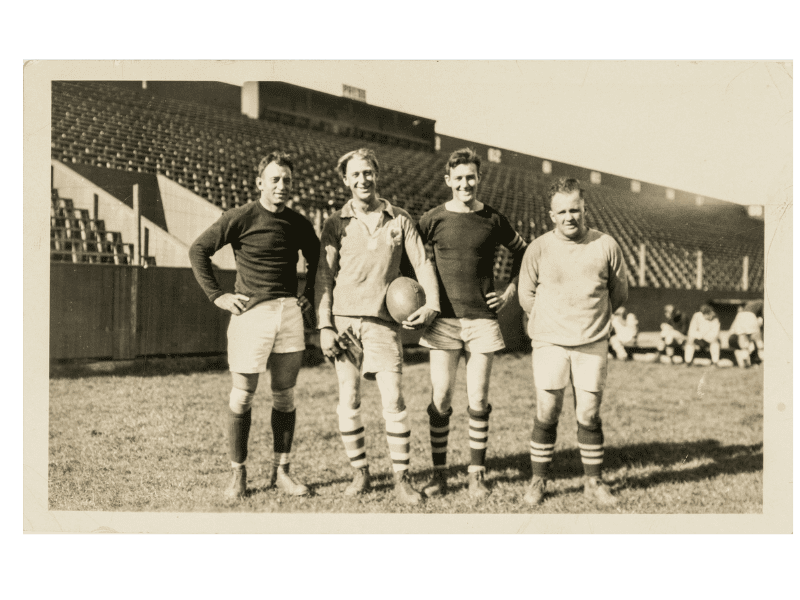Black-and-white photo of four rugby players standing side-by-side on a field in front of empty bleachers. The second from left holds a rugby ball under one arm.