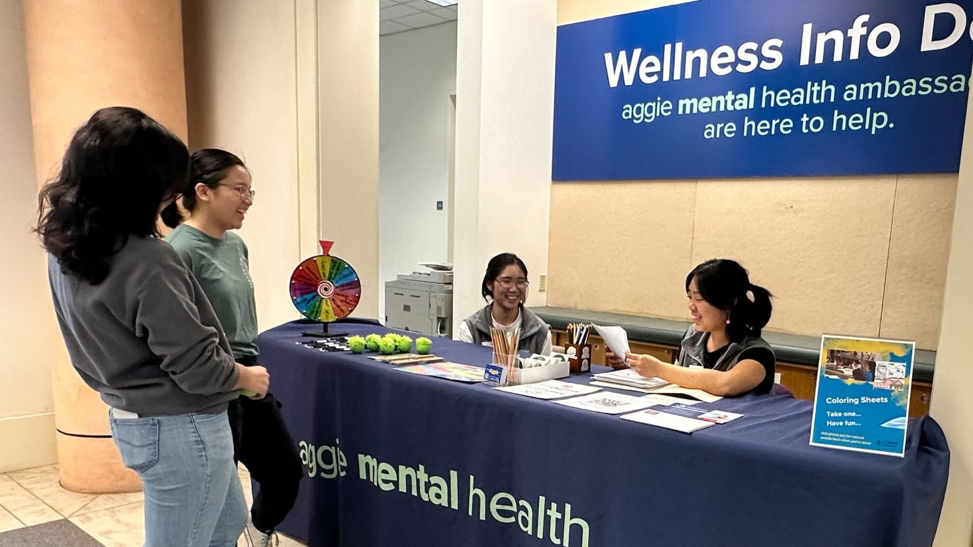 Two students sit behind a desk draped with a blue Aggie Mental Health tablecloth. Flyers and a spin-the-wheel game are on the desk. Two other students stand in front of the desk. All four students are smiling.