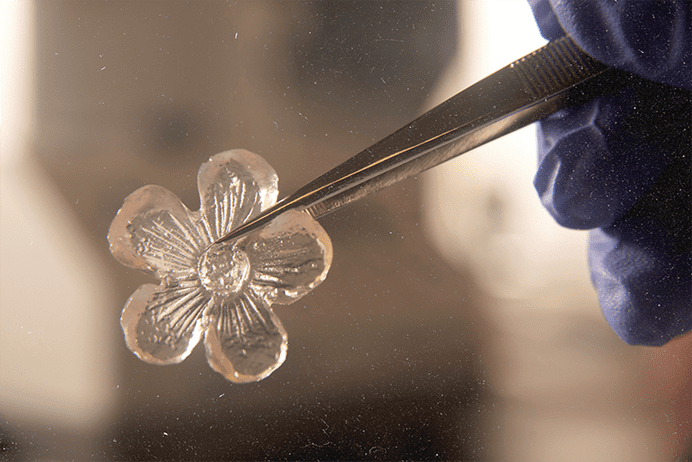 Close up of flower-shaped jelly ice cube held by tweezers and a gloved hand.