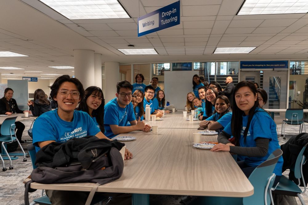 A group of student peer tutors in blue Academic Assistance and Tutoring Center t-shirts sitting together at a table smile for the camera