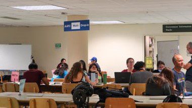 Students studying in a brightly lit space with old 1960s-style wooden chairs. A blue sign for Math tutoring hangs from the ceiling.