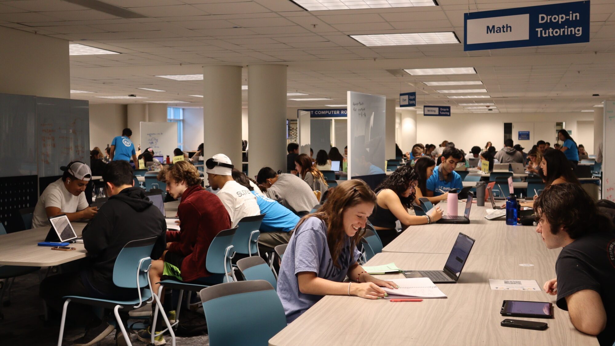 Smiling students study in a brightly lit space with modern tables and chairs. A blue sign for Math tutoring hangs from the ceiling.