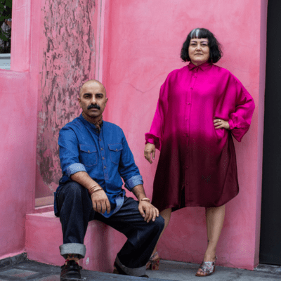 Against a hot pink background, a person seated, wearing a denim shirt and cuffed jeans next to a person standing wearing a hot pink blouse and black skirt.