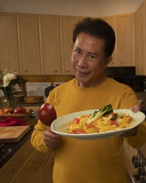 Chef Martin Yan smiles, holding out a plate of stir fry. A cutting board and basket of vegetables are in the background.