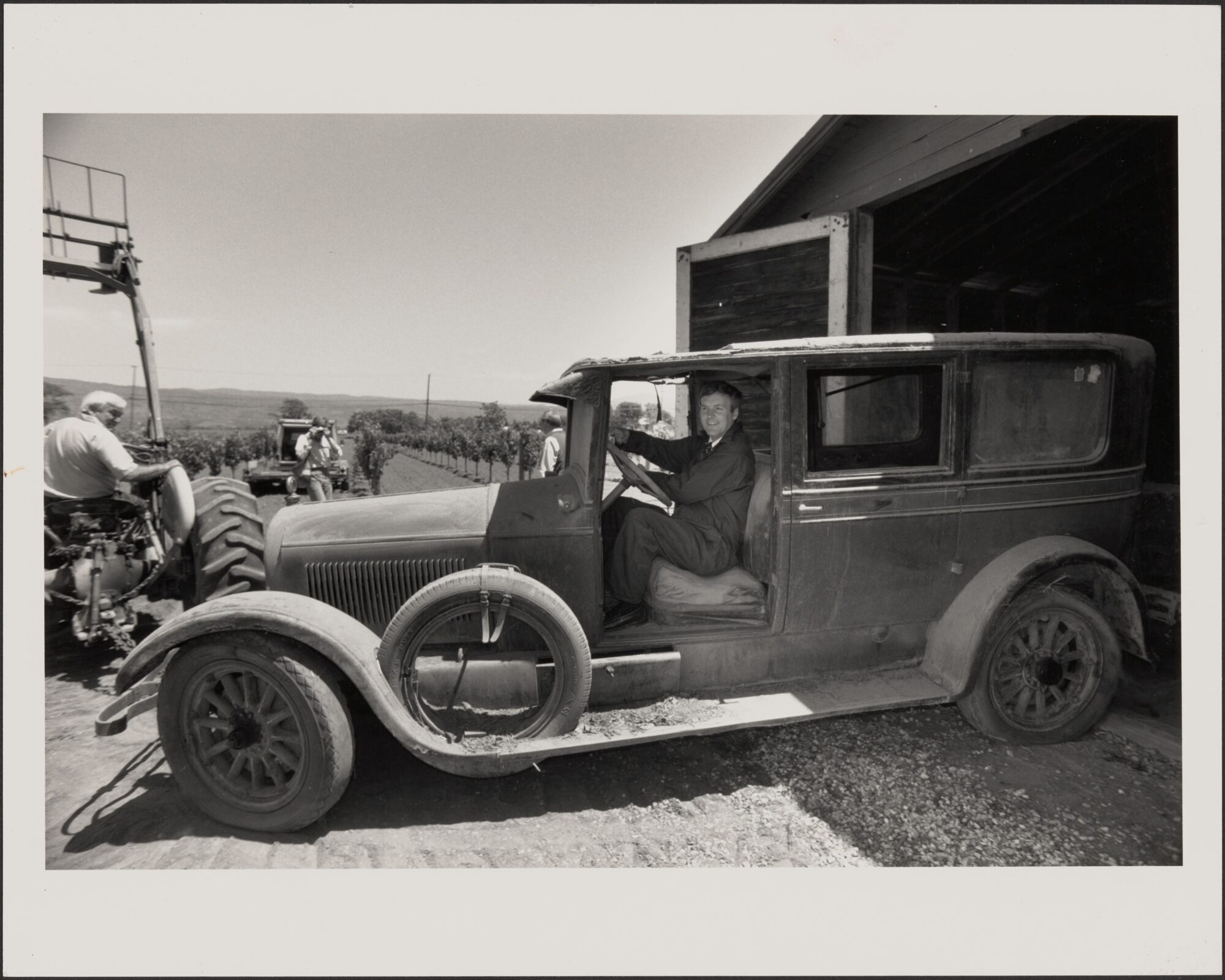 Black and white image of Cadillac parked in barn entrance with man in driver's seat and tractor and vineyard in background.
