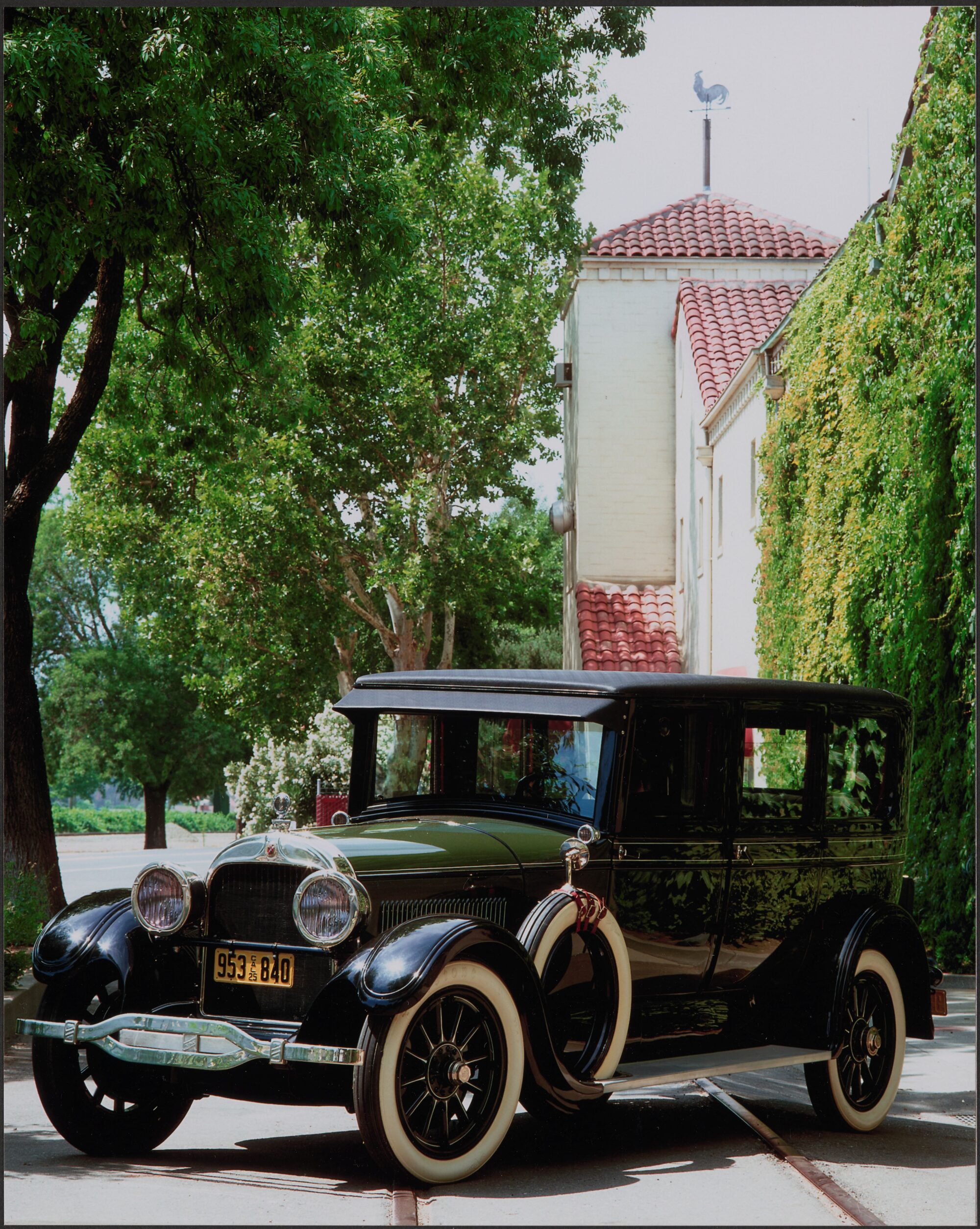 Black restored Cadillac parked in front of BV with trees on left and vine-covered wall on right.