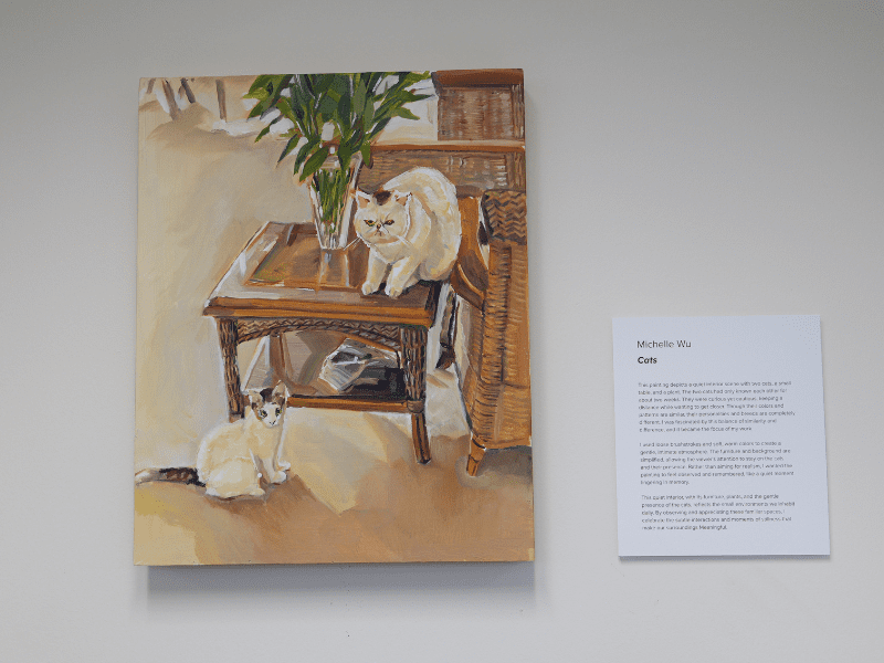 Two cats stare at viewer one on floor and one on top of side table next to vase filled with green stems.