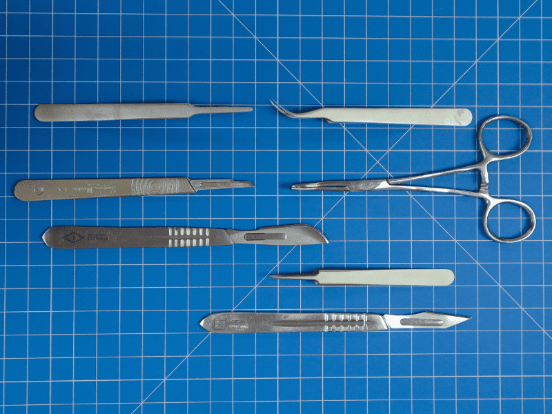 Three scalpels and four precision tweezers of varying size and shape on a blue gridded matt