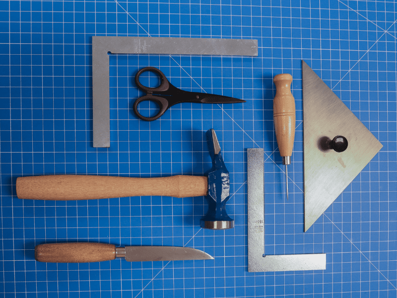 Bookbinders tools arranged on a blue gridded mat, including two rules, an awl, hammer and precision scissors.