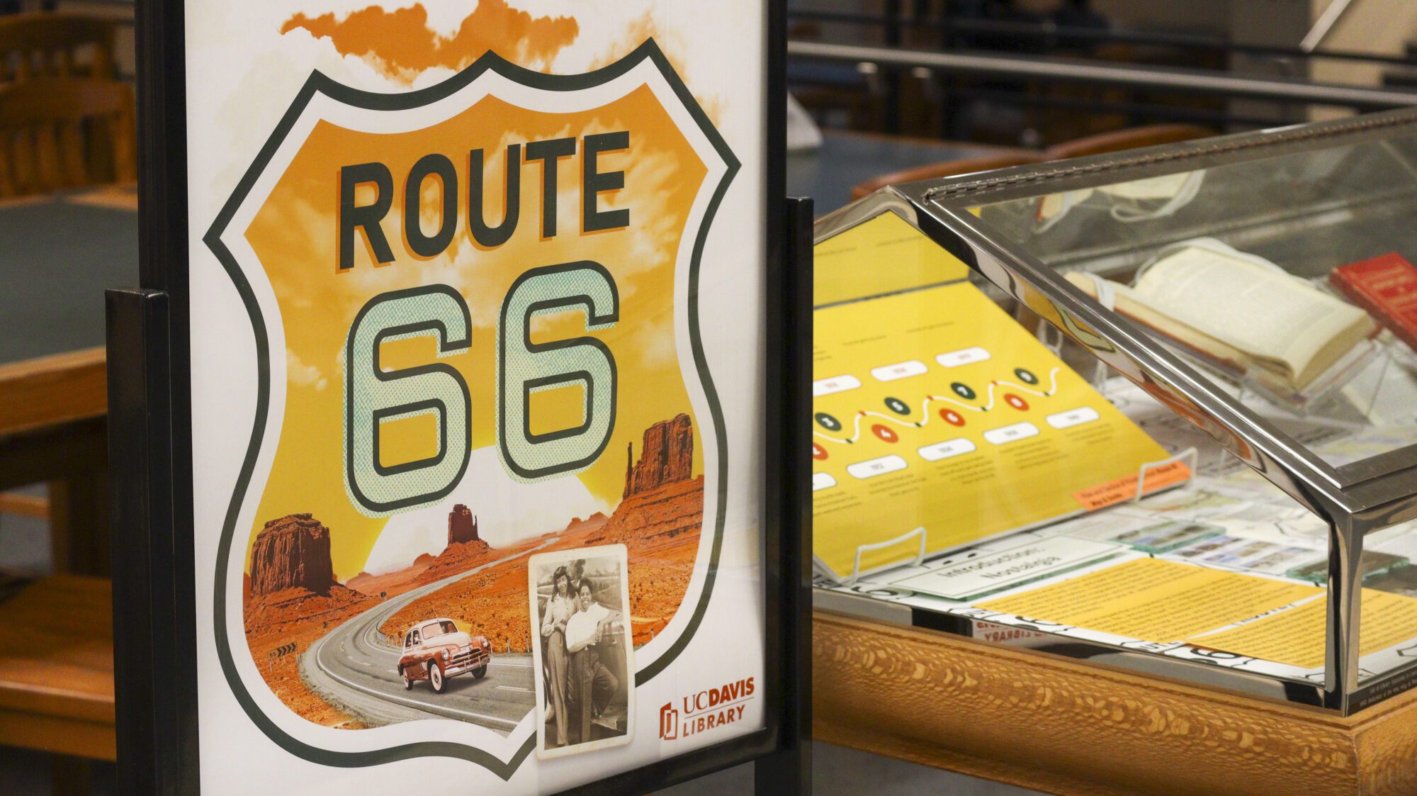 Route 66 exhibit display with a poster showing a desert highway and Route 66 shield beside a library exhibit case containing historical travel materials.