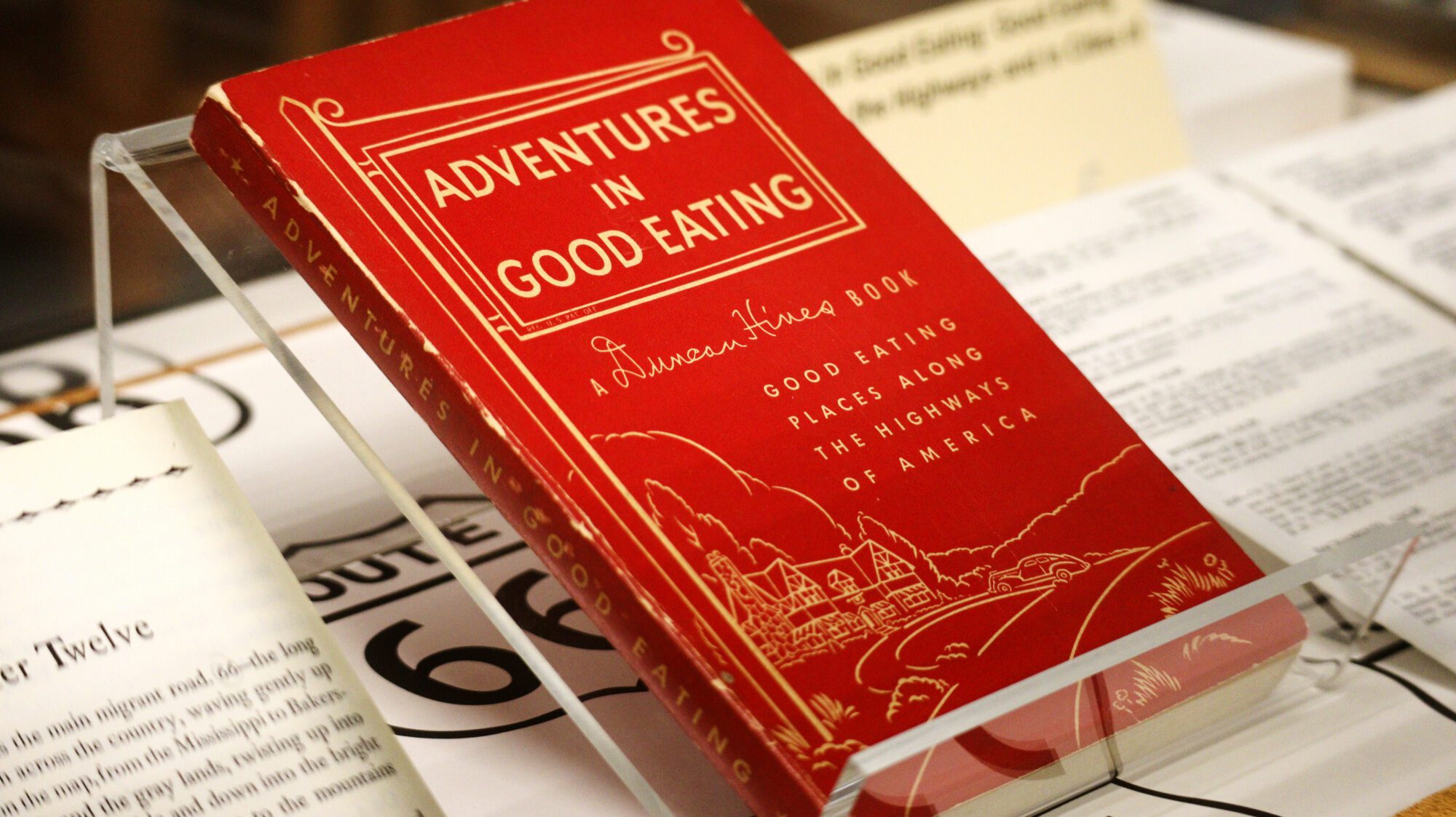 Red cover of the vintage guidebook Adventures in Good Eating displayed in a Route 66 exhibit case.
