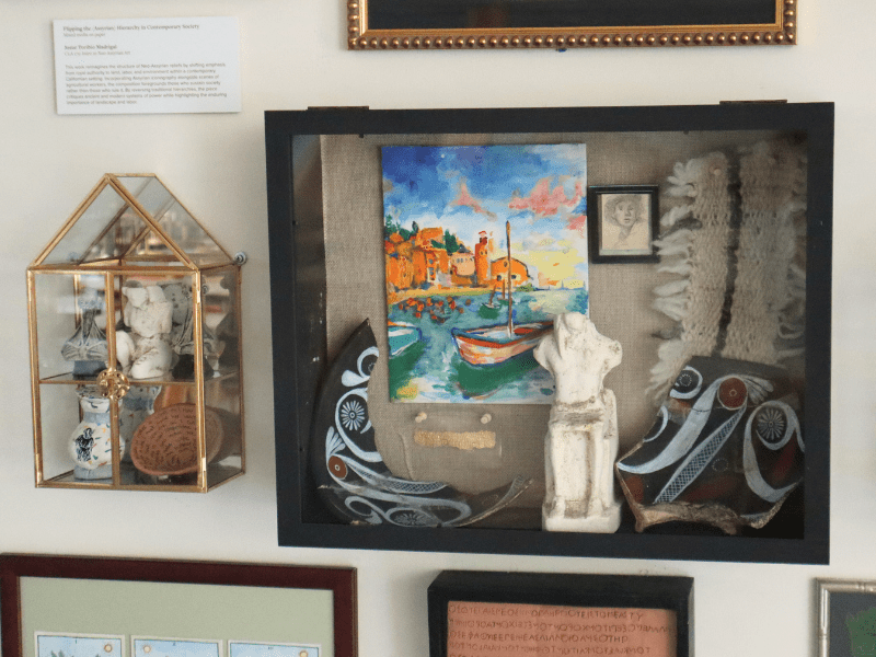 Framed art works including mixed media in miniature copper-colored houses and a shadow box with a painted canvas of a boat and a plaster figure, displayed on a wall with captions