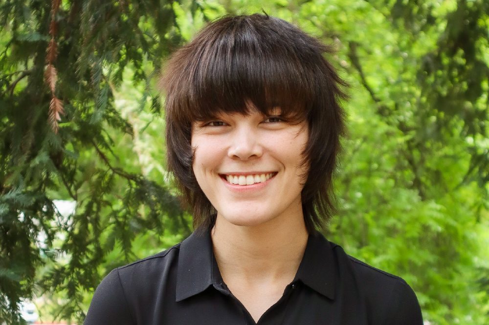 Alyssa Linehan poses wearing a black collared shirt, with green trees in the background
