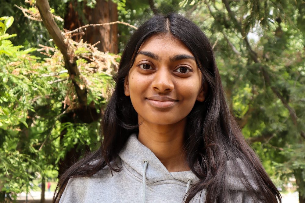 Portrait of Ruth Selvaraj with green treen branches in background.

