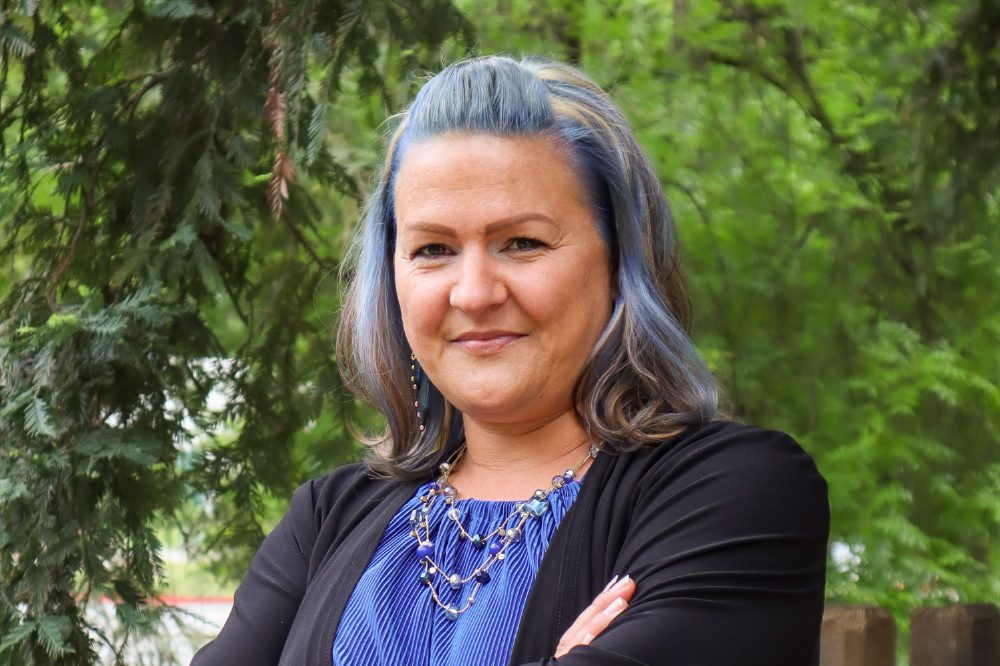 Sofia Wright poses wearing a black cardigan over a blue shirt, with green trees in the background