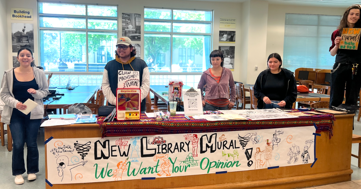 Four students stand behind a large wood table decorated with a colorful poster titled "New Library Mural We Want Your Opinion! One students stands to the right on a chair holding a sign "NEW mural help us out!"