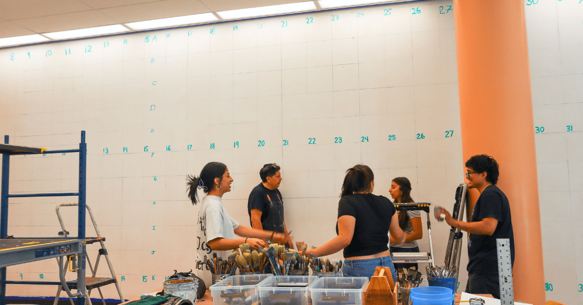 Students gather around their instructor in front of a gridded wall, labeled with numbers and letters. Buckets and clear containers of painting supplies are in the foreground, with scaffolding and step ladder to the left.