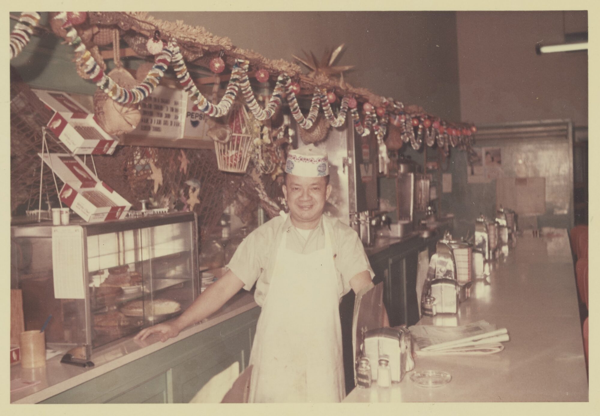 A man in a white chef's apron stands, smiling, behind a diner-style restaurant counter.