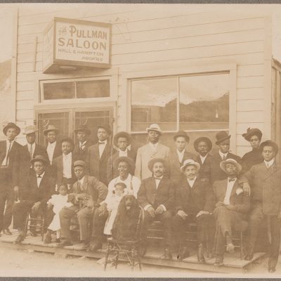 This photograph is a group portrait of African American men, women, and children posed outside the Pullman Saloon in Tonopah, Nevada, circa 1905. The image contains eighteen men, one woman, two young girls, and two dogs. Several of the men are smoking cigars. A spaniel is seated on a chair in front of the group.