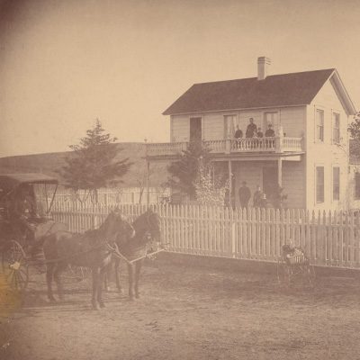 Portion of the photo which shows a farmhouse with seven people standing on both the upstairs and downstairs porches. A horse, buggy, and baby carriage appear on the road in front of the house.
