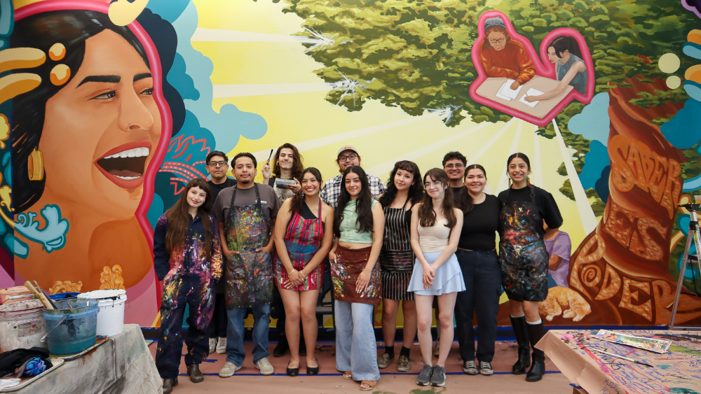 Student artists pose in front of a large, colorful wall-to-wall mural that includes a grito, oak tree, students in various daily life and other elements.