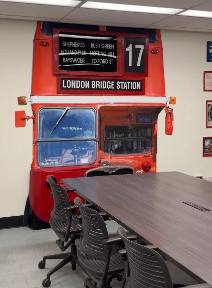A conference-style table with three chairs in the foreground. Beyond it, on the wall, is the front of a red Unitrans bus made up partly of a life-size photograph and partly of actual bus parts, including rearview mirrors, front fender and destination sign bearing original London locations.