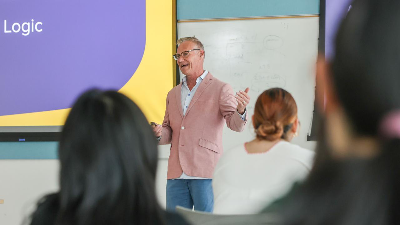 A man wearing glasses and a blazer lectures to a group of students