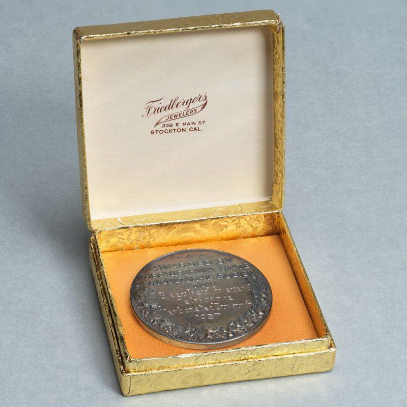 A silver medal in a gold-foil wrapped jewelers box. The box bears the logo of Friedbergers Jewelers in Stockton, Cal.
