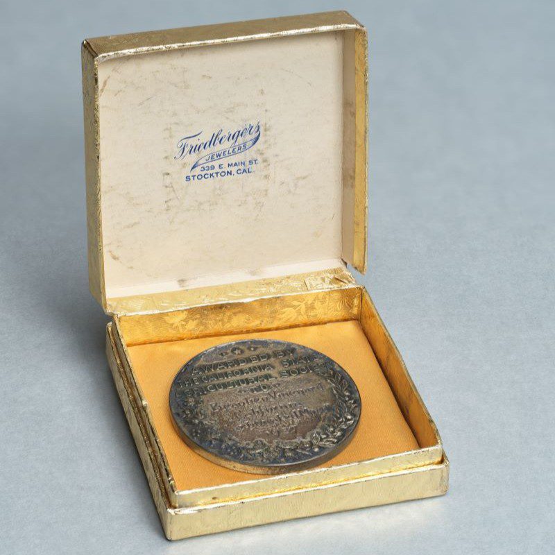 A silver medal in a gold-foil wrapped jewelers box. The box bears the logo of Friedbergers Jewelers in Stockton, Cal.
