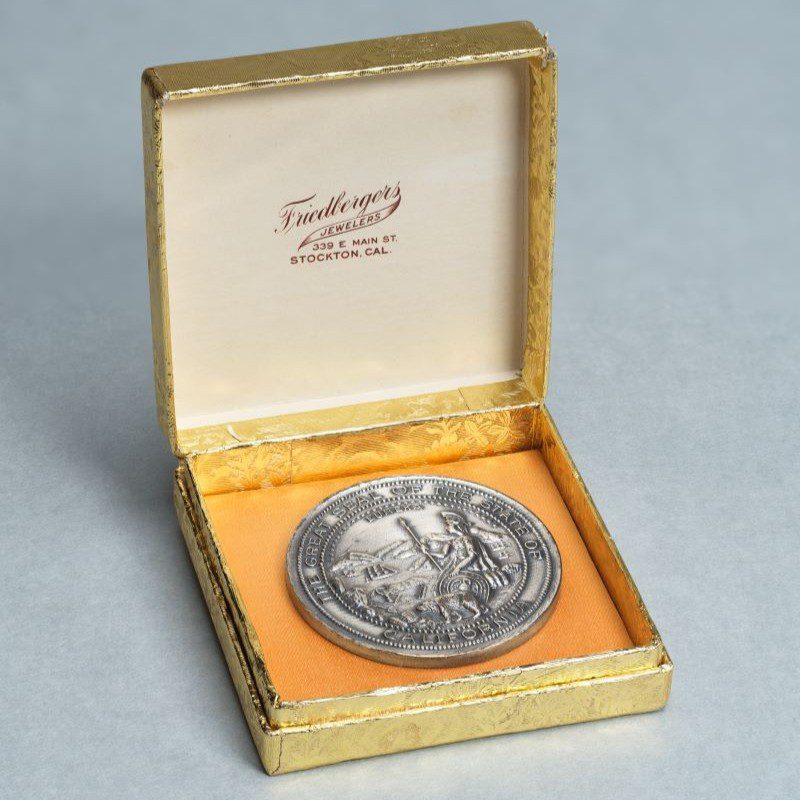 A silver medal embossed with the Great Seal of the State of California, displayed in a gold-foil wrapped jewelers box.