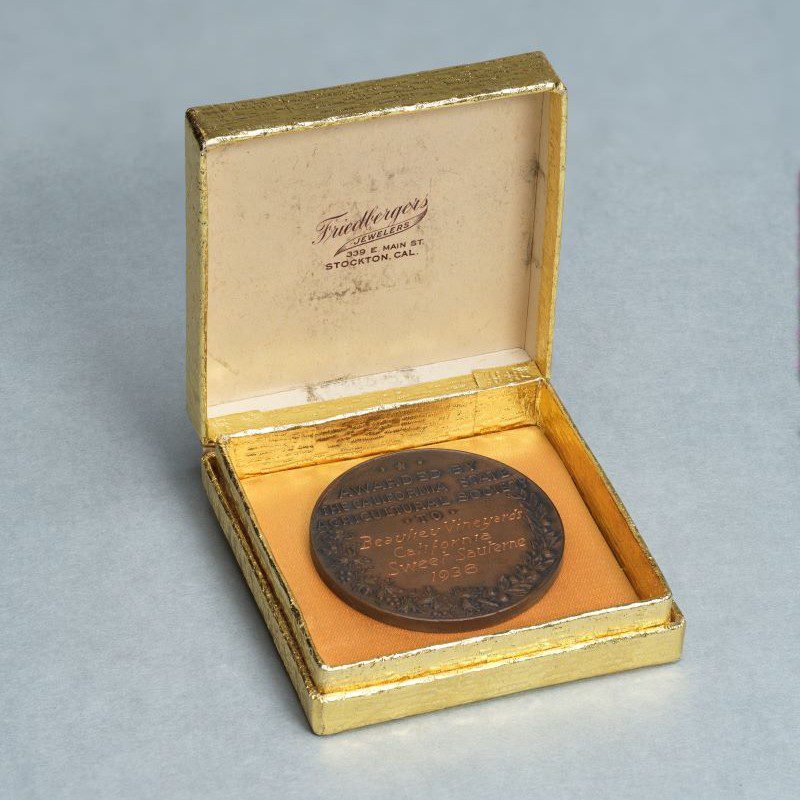 A bronze medal in a gold-foil wrapped jewelers box. The box bears the logo of Friedbergers Jewelers in Stockton, Cal.