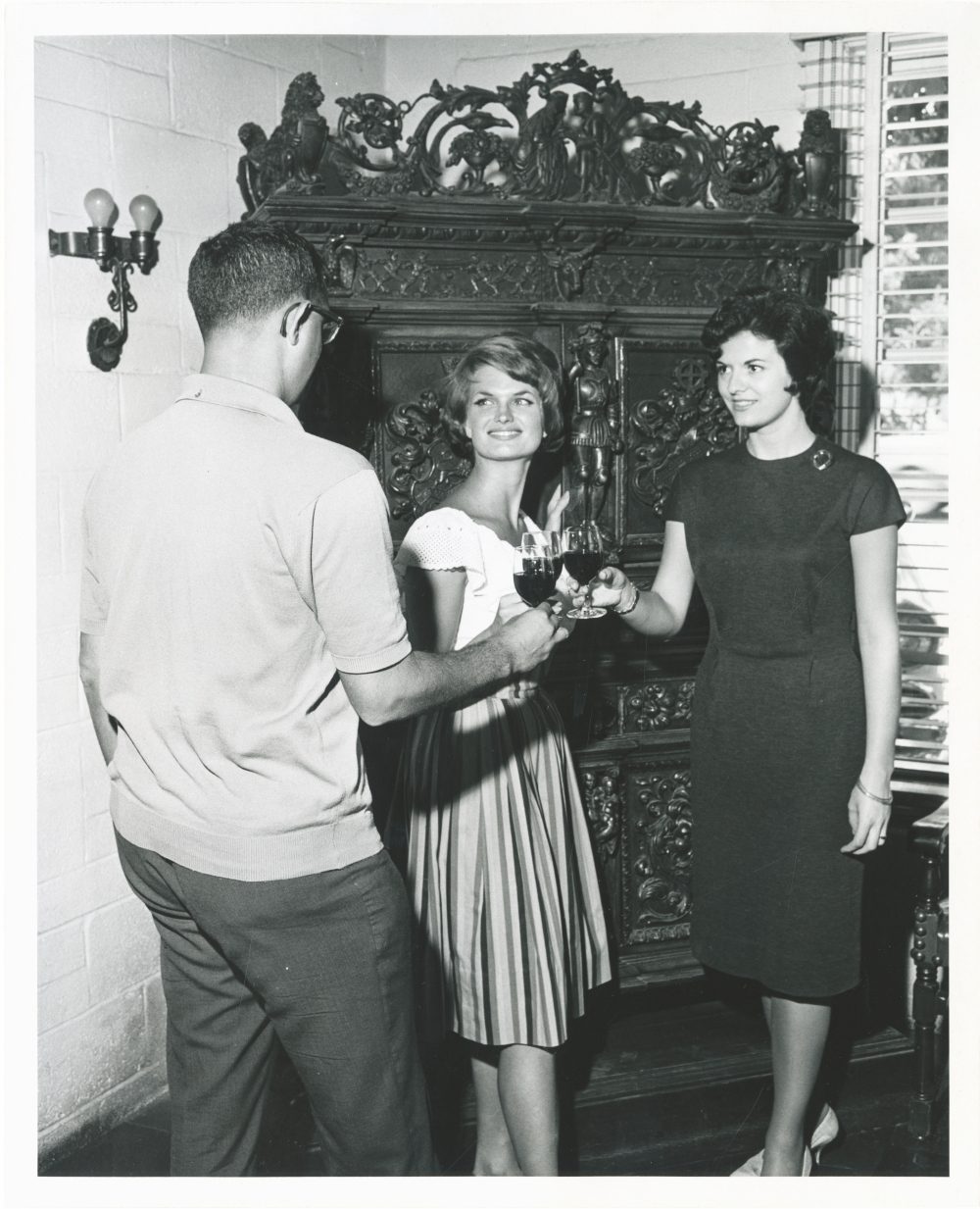 One man and two women in 1960s era clothing clink glasses. An ornate wood cabinet is in the background behind them.