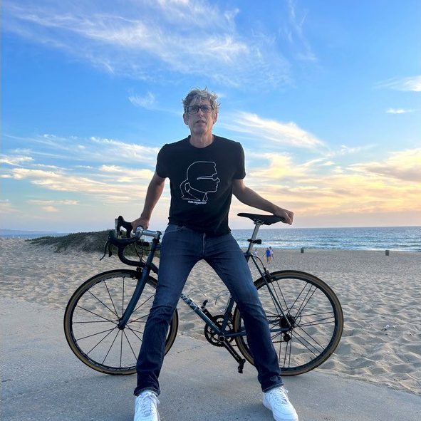 Peter Flax wearing glasses stands in front of a bicycle. A blue sky, sand, and water are in the background.