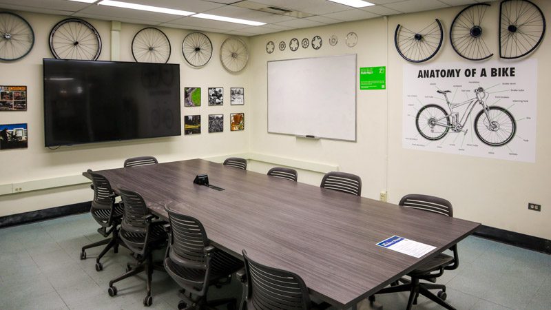Conference table with mounted bicycle wheels lining the upper walls and an 'Anatomy of a Bike' diagram poster, with a mounted TV screen and whiteboard.