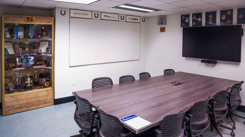 Conference table across from a rustic wood display cabinet holding trophies and awards, with a mounted TV screen and whiteboard.