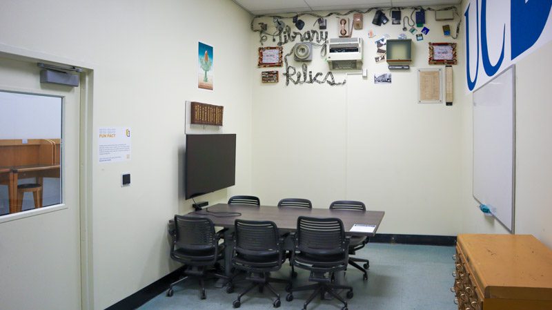 Conference table and chairs in a room with a wall display of vintage library photos, small artifacts and 'Library Relics' lettering, plus a mounted TV screen.